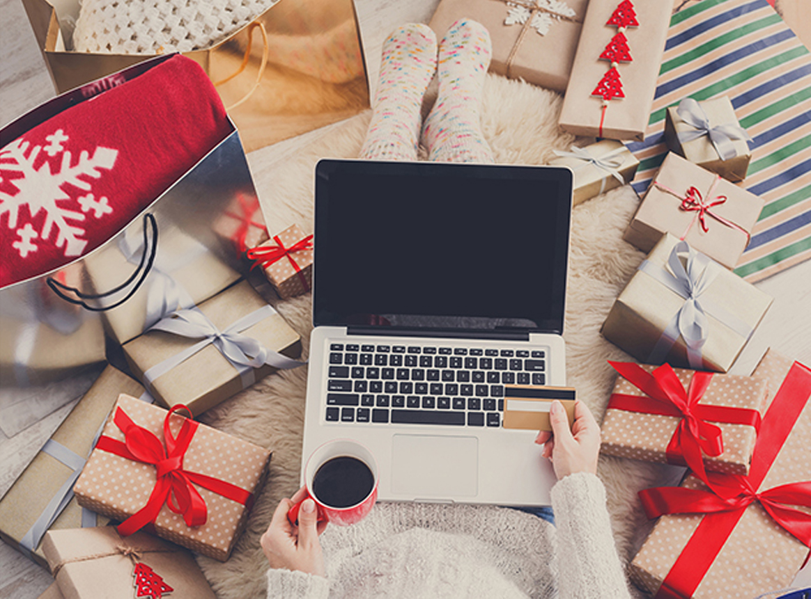 Woman sitting, surrounded by holiday presents, with laptop on her lap and shopping online